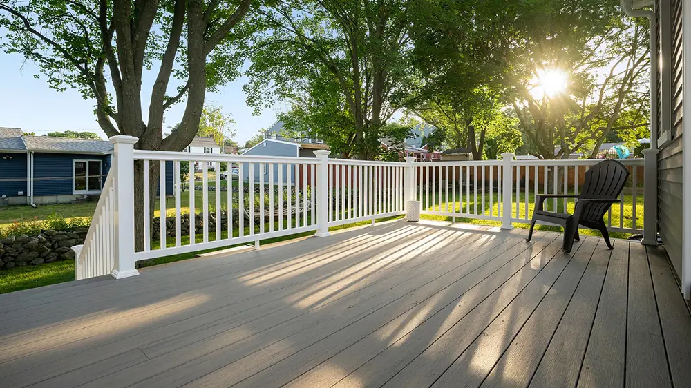Sunlit wooden deck with white railing and a single black chair, overlooking a suburban yard with trees.